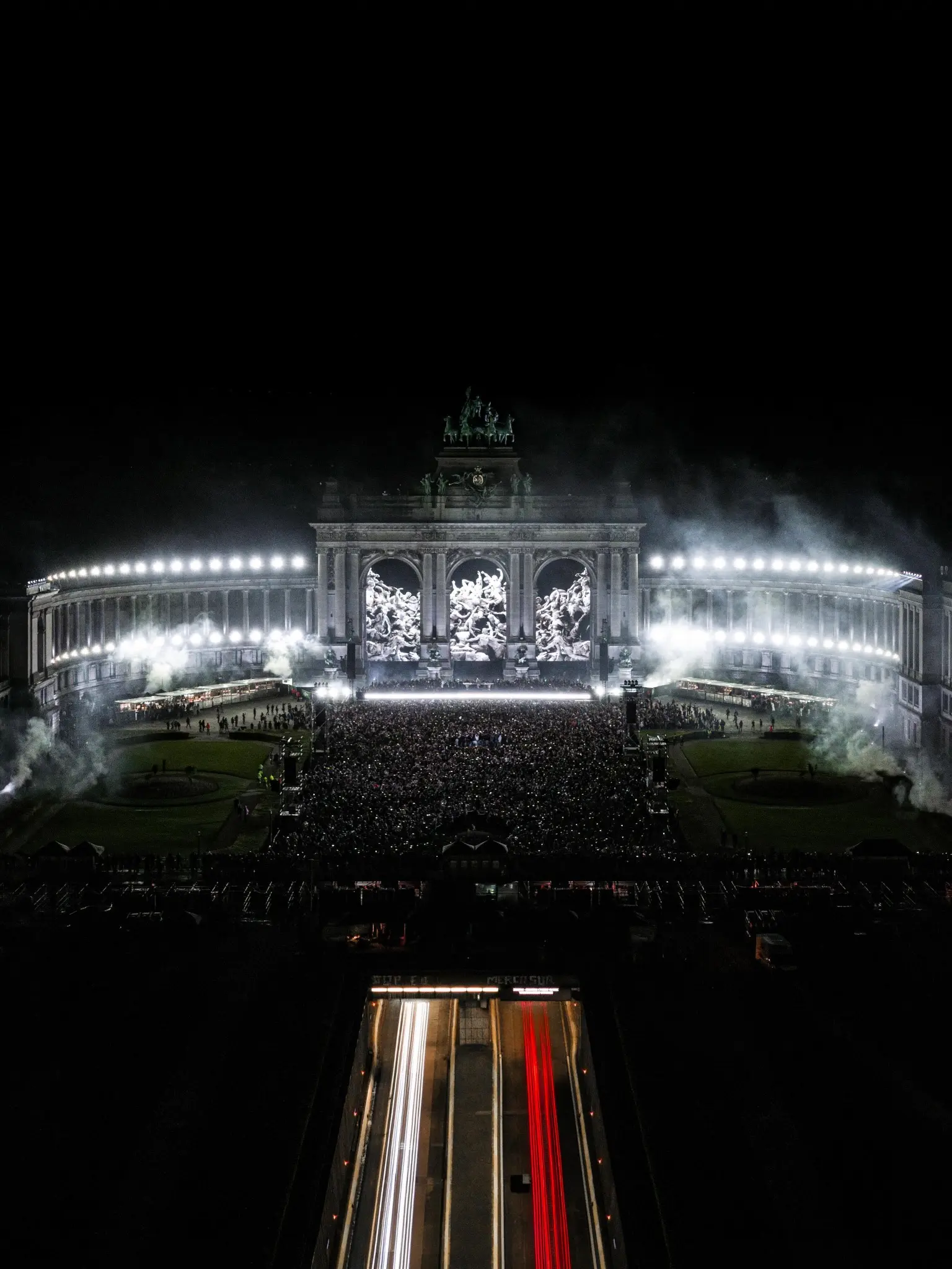 A packed crowd at night in front of the lit colonnade of the Cinquantenaire in Brussels, seen from above