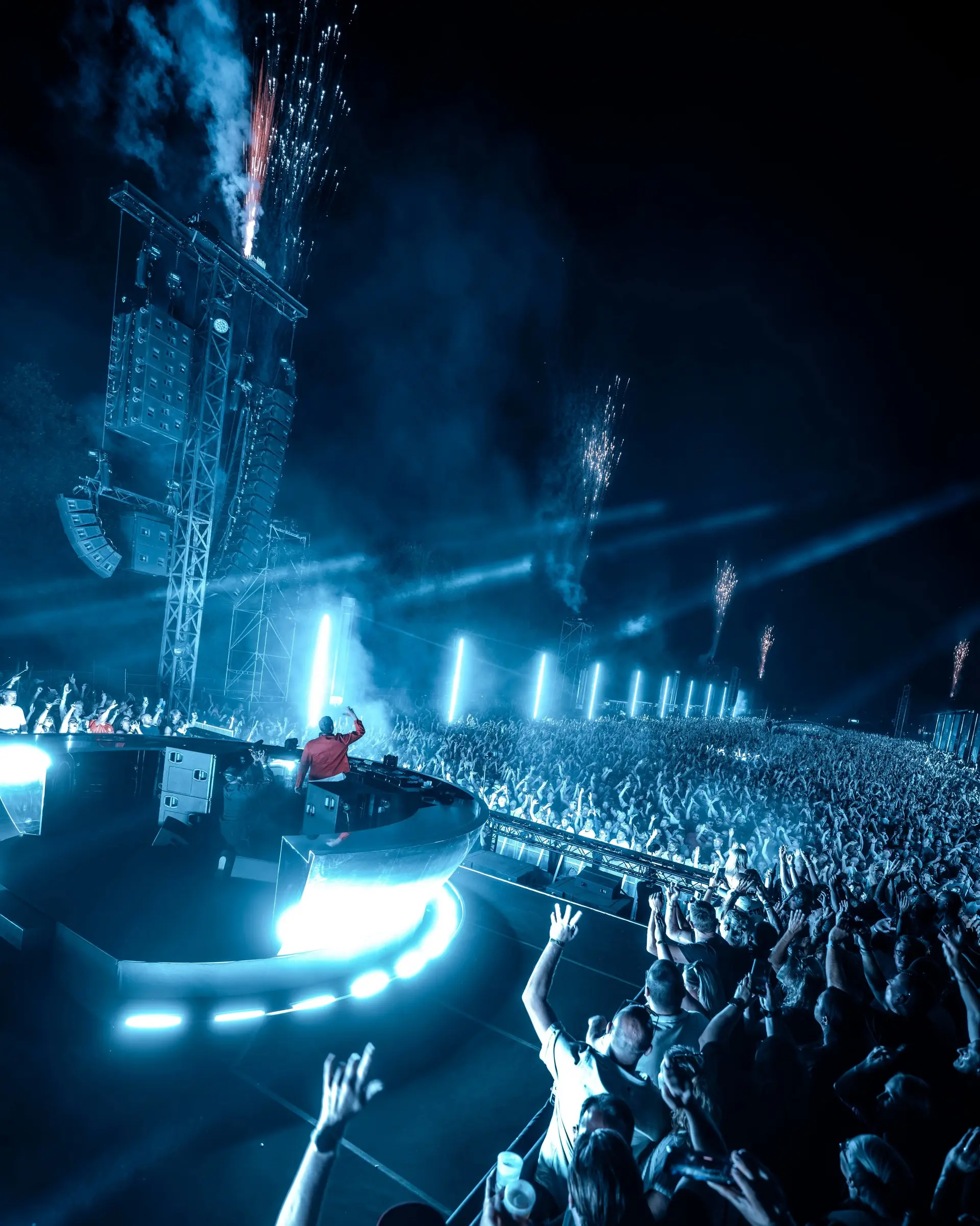 A DJ on a lit circular stage above a huge crowd with raised hands, fireworks over the field