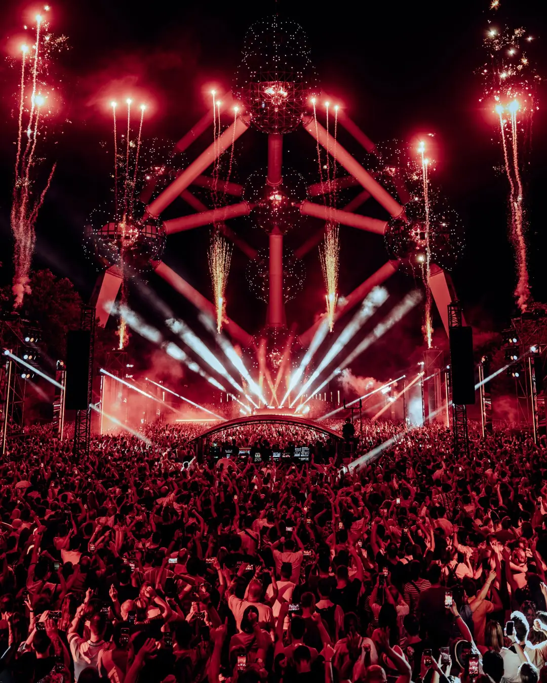 A packed crowd in red light in front of the Atomium, with fireworks rising from the stage