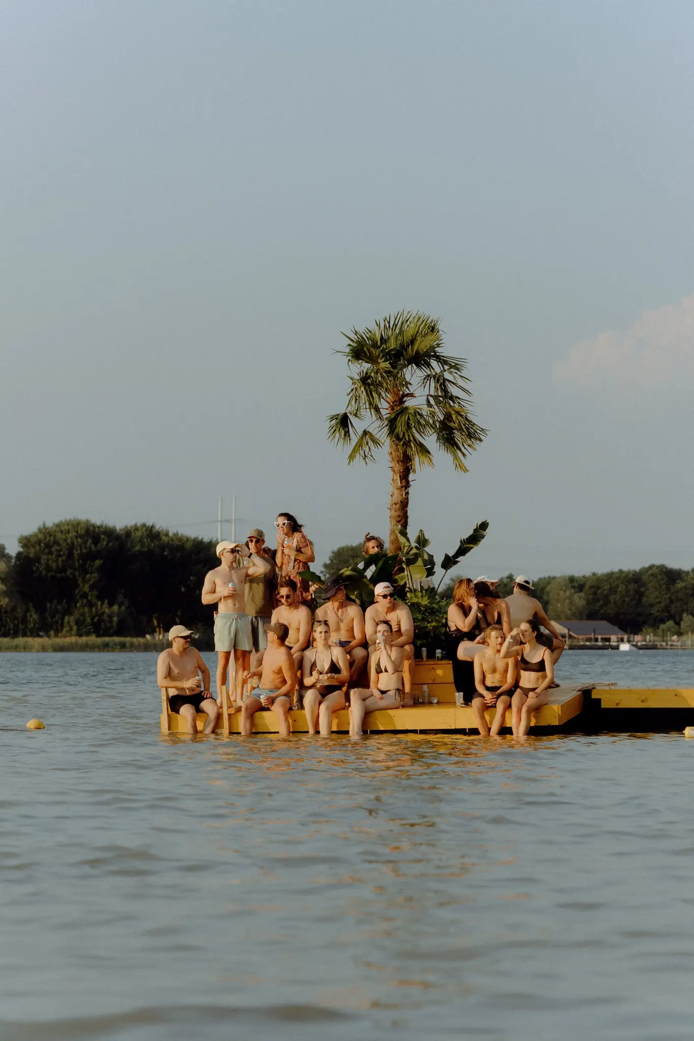 Festivalgoers on a floating pontoon with a palm tree on the lake at Lago Lago